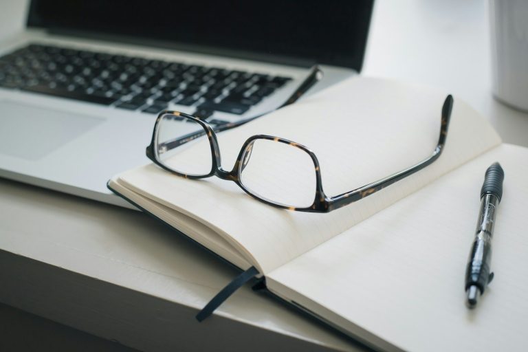 Laptop, Brille, ein leeres Notizbuch und ein Stift auf einem Tisch.