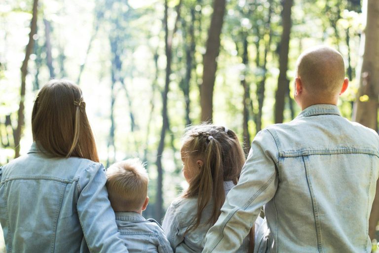 Familie aus vier Personen schaut nach vorne in einen Wald. Sonnenlicht durchbricht die Bäume.