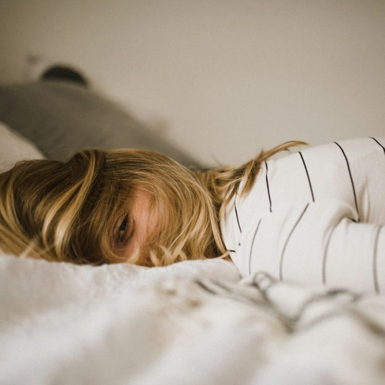 Eine Person liegt erschöpft mit dem Gesicht nach unten auf einem Bett, umgeben von Kissen.