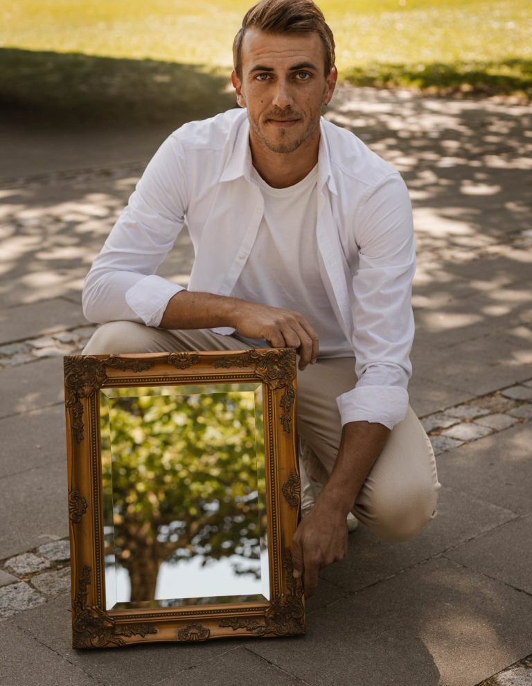 Benjamin Feurer kniet neben einem goldenen Spiegelrahmen mit reflektierendem Baum.