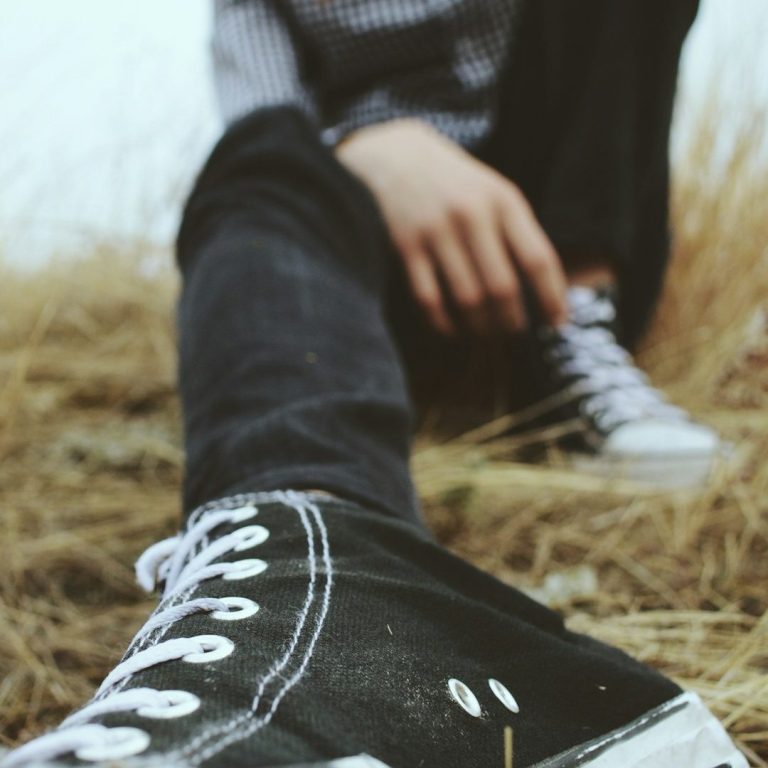 Schwarze Sneakers auf einem mit trockenem Gras bedeckten Boden, Jugendlicher im Hintergrund.